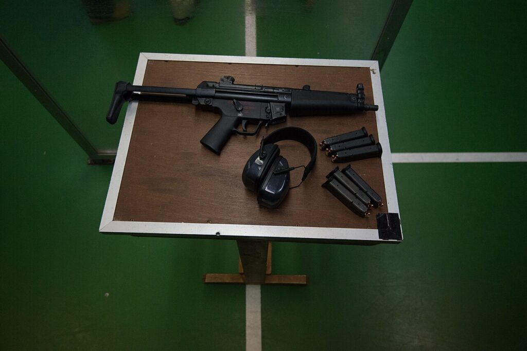 A firearm, hearing protection earmuffs, and magazines arranged on a table at an indoor shooting range.