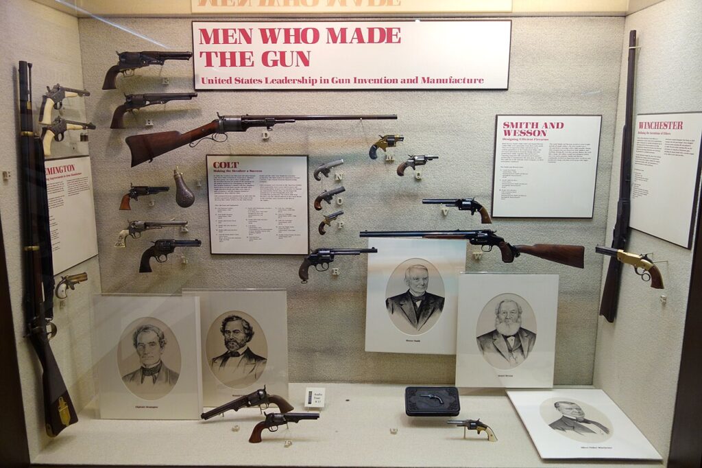 A museum display case containing various historical firearms, portraits of inventors, and informational plaques about gun manufacturing history.