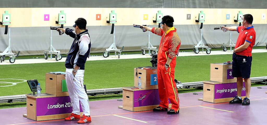 Three competitive shooters in uniform stand at a firing line, aiming pistols at targets during an international competition.