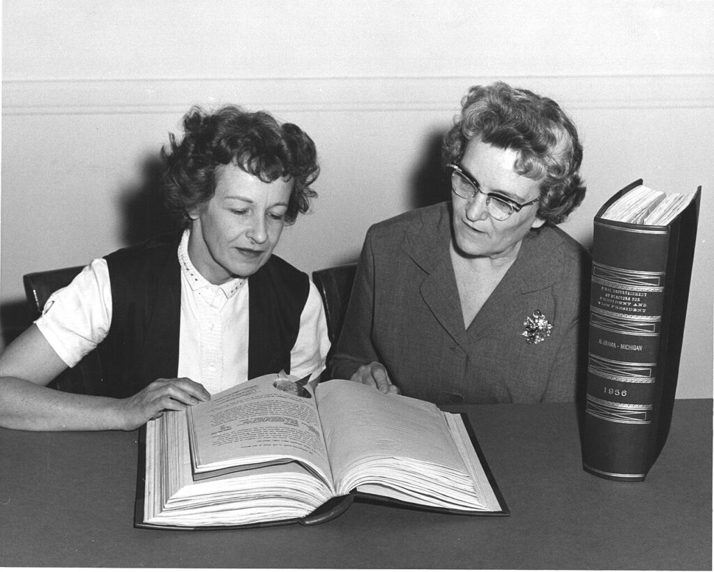 Two women reviewing and examining thick, official government records and documents at a table.