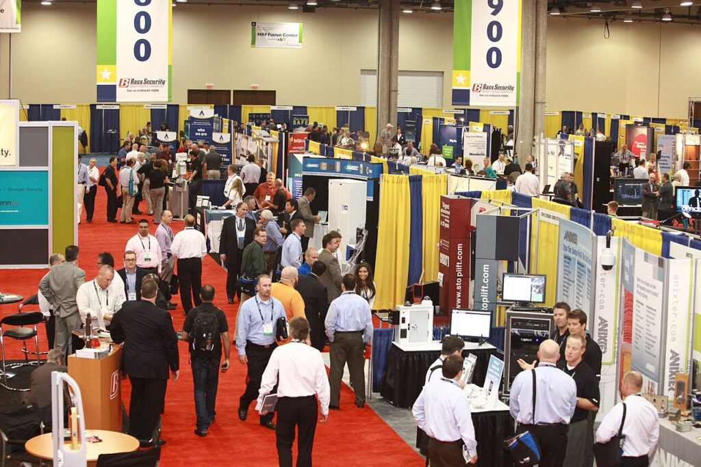 Attendees walking through a large, busy trade show floor with various exhibitor booths and banners.