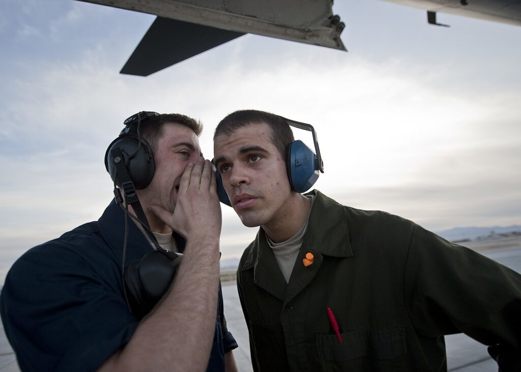 Two people wearing protective earmuffs while one whispers to the other on an airfield.