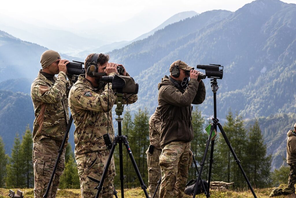 Soldiers using spotting scopes on tripods in a mountainous, outdoor environment during the daytime.