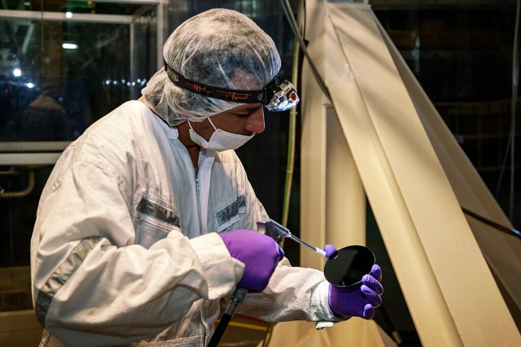 A technician wearing protective gear carefully cleans a circular optical component with a specialized tool.