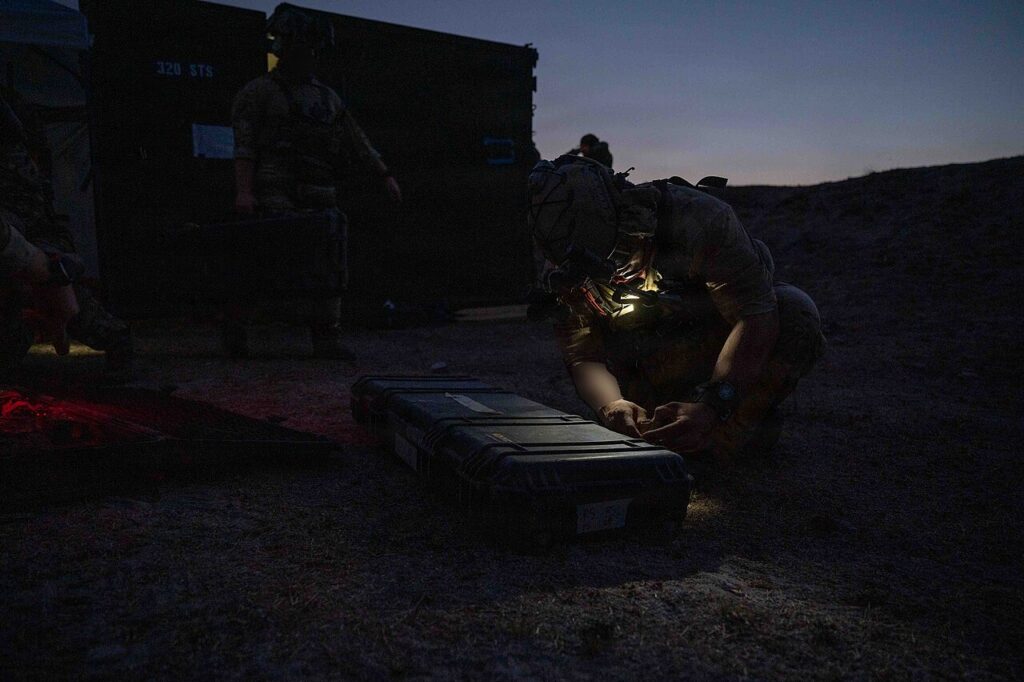 A soldier in dim lighting kneels to inspect and prepare a black, hard-shelled protective equipment case.