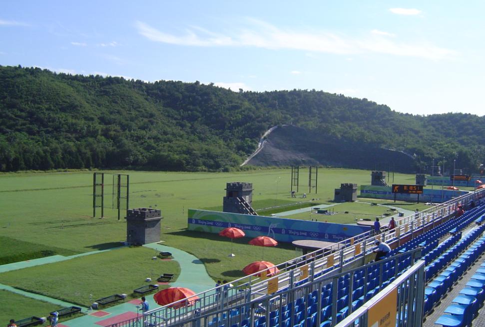 A wide outdoor shooting range with blue stadium seating under a clear sky.