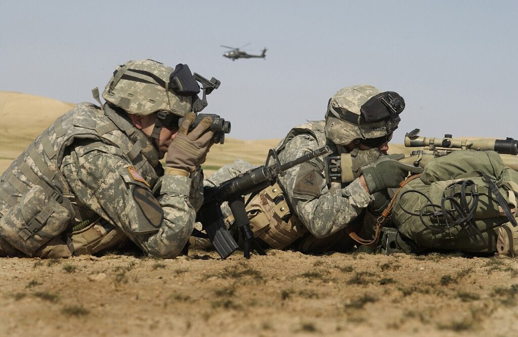Two soldiers in camouflage uniforms lying prone while using binoculars and a riflescope in a field environment.