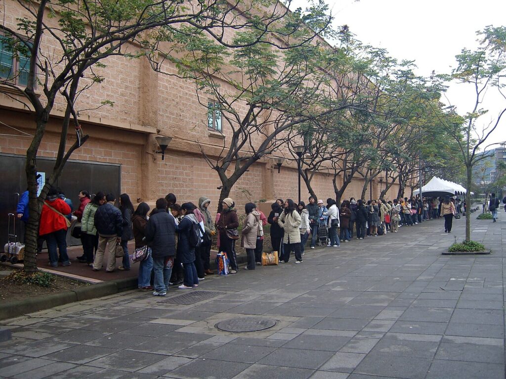A long line of people waiting outside a large exhibition hall building before an event opens.