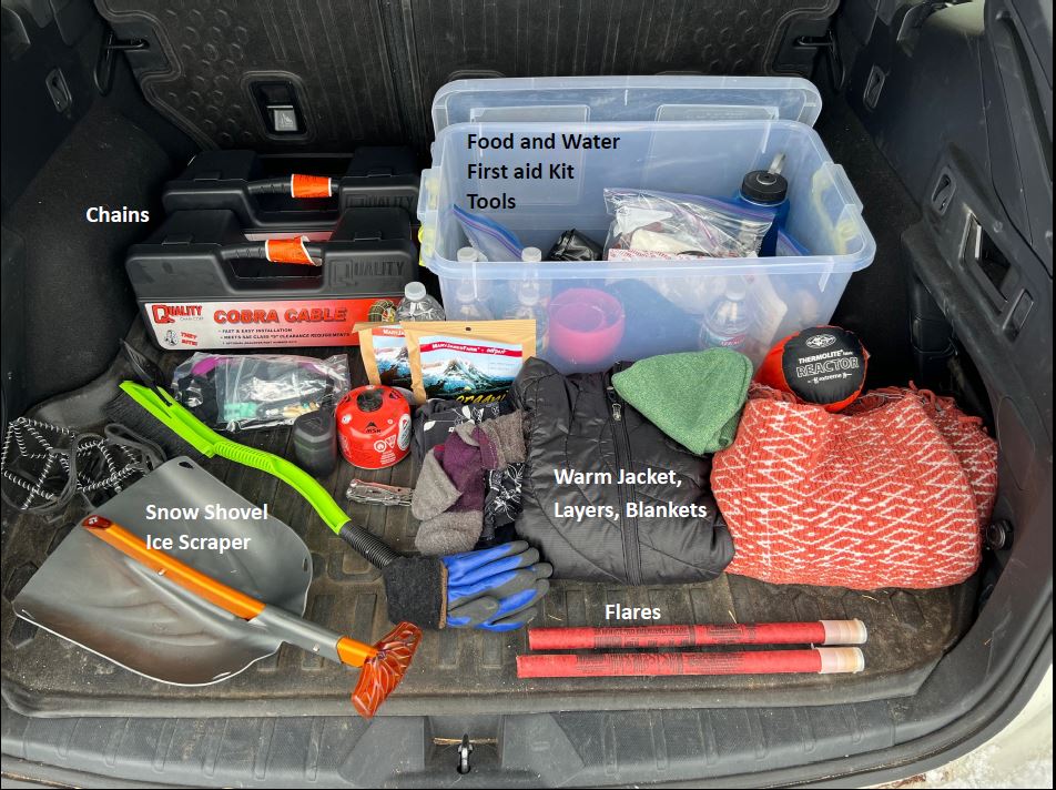An open car trunk containing a plastic bin of emergency supplies, a snow shovel, and warm winter clothing.
